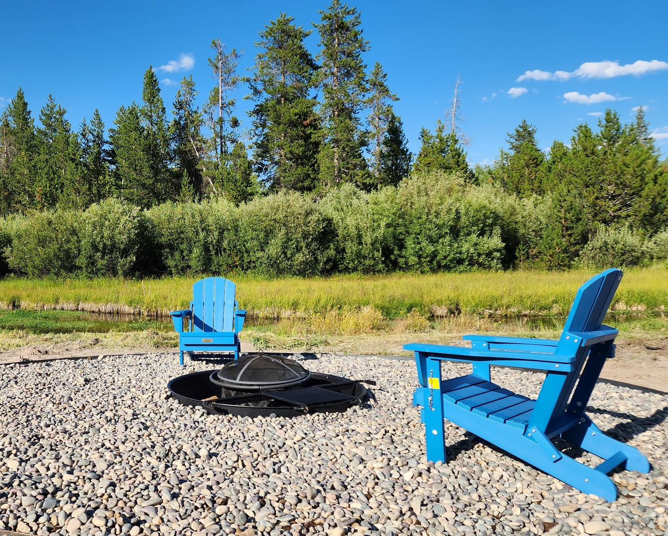 Two blue Adirondack chairs sit by a fire pit on a gravel area; trees and a blue sky in the background.