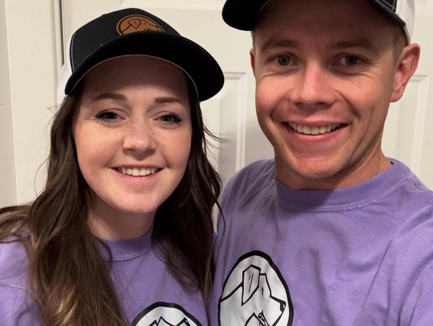 A man and a woman wearing purple shirts and hats are smiling for a picture.