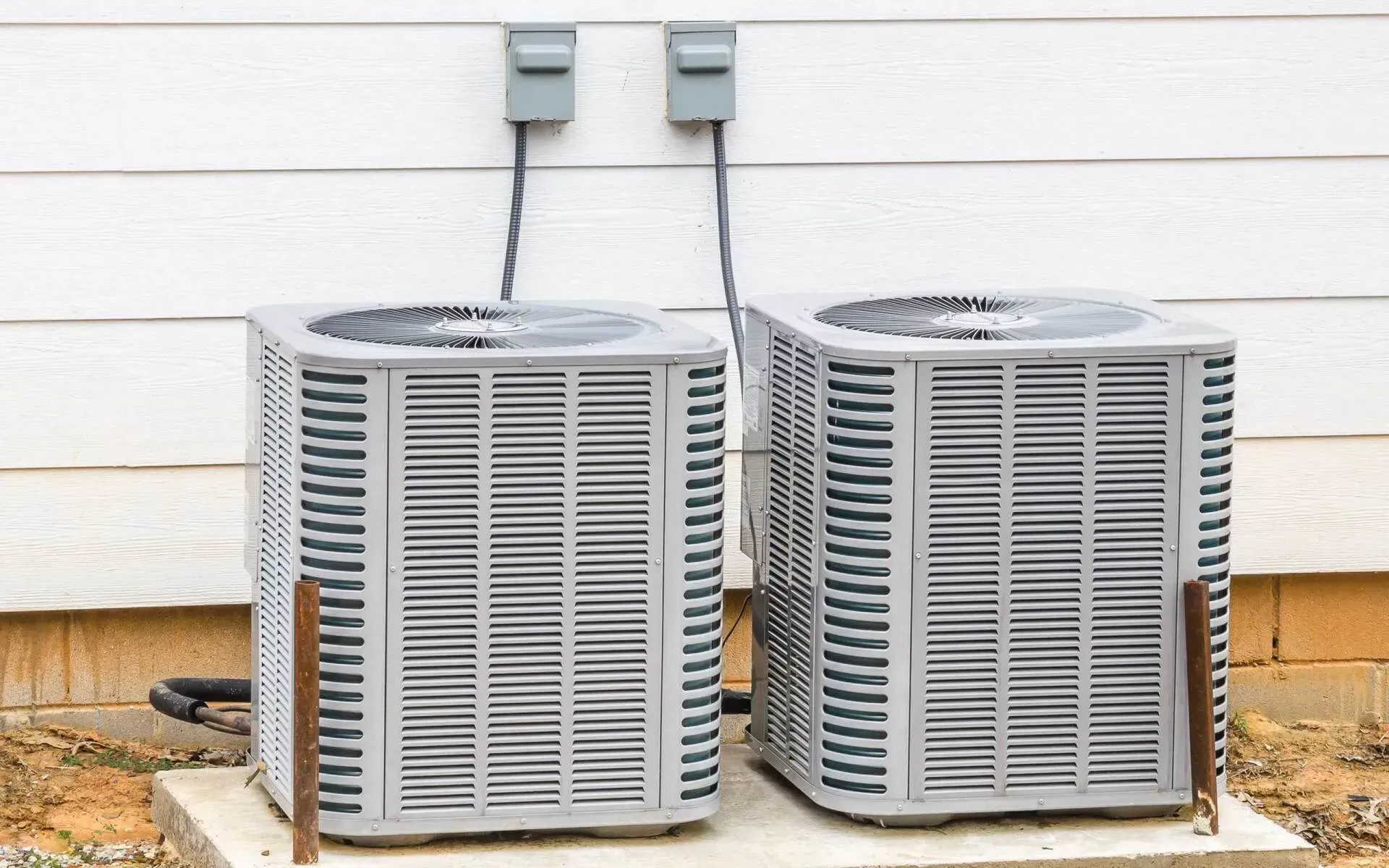 Two air conditioning units next to a white house, connected to electrical boxes above.