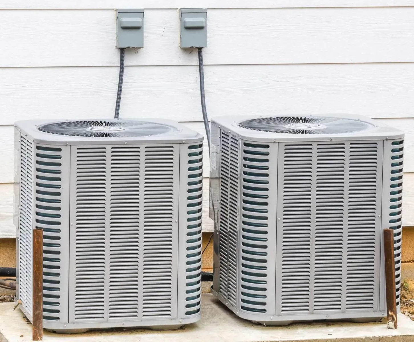 Two air conditioning units next to a white house, connected to electrical boxes above.
