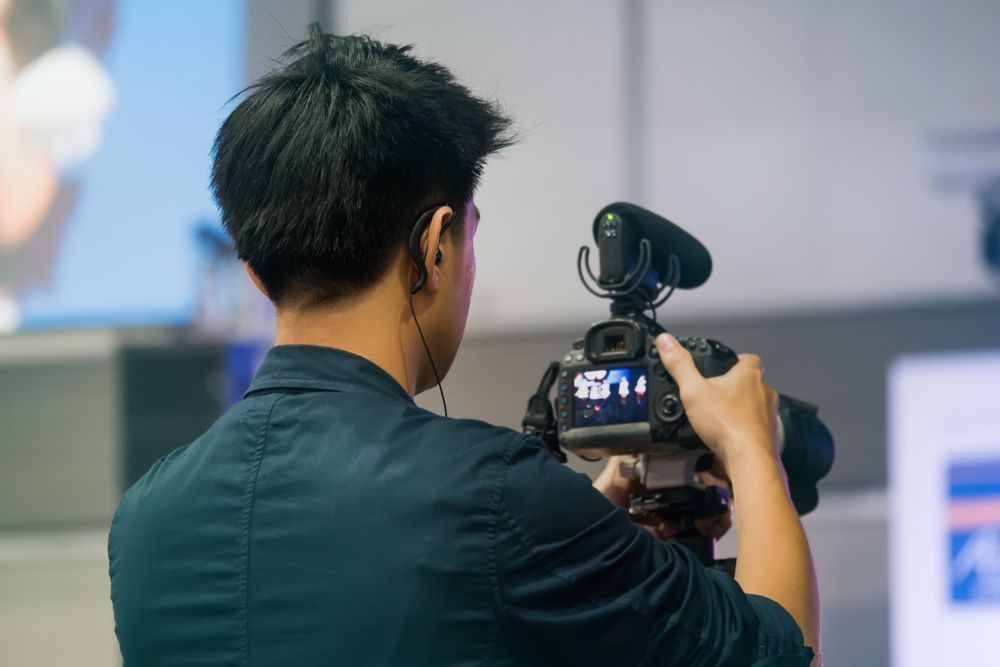 Videographer filming event, holding camera with attached microphone, focused on a screen in the background.