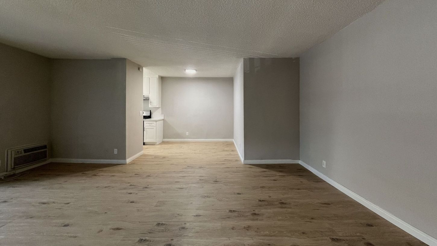 Empty apartment interior with gray walls, wood-look flooring, and a small kitchen visible in the back.