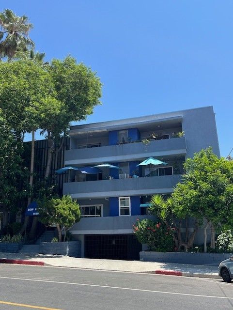 Three-story apartment building with blue accents and umbrellas on balconies, street view under a clear sky.