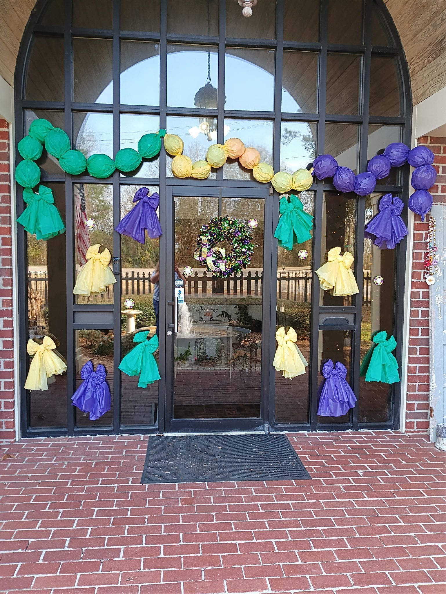 A glass entrance door decorated with green, yellow, and purple tissue paper pom-poms for Mardi Gras.