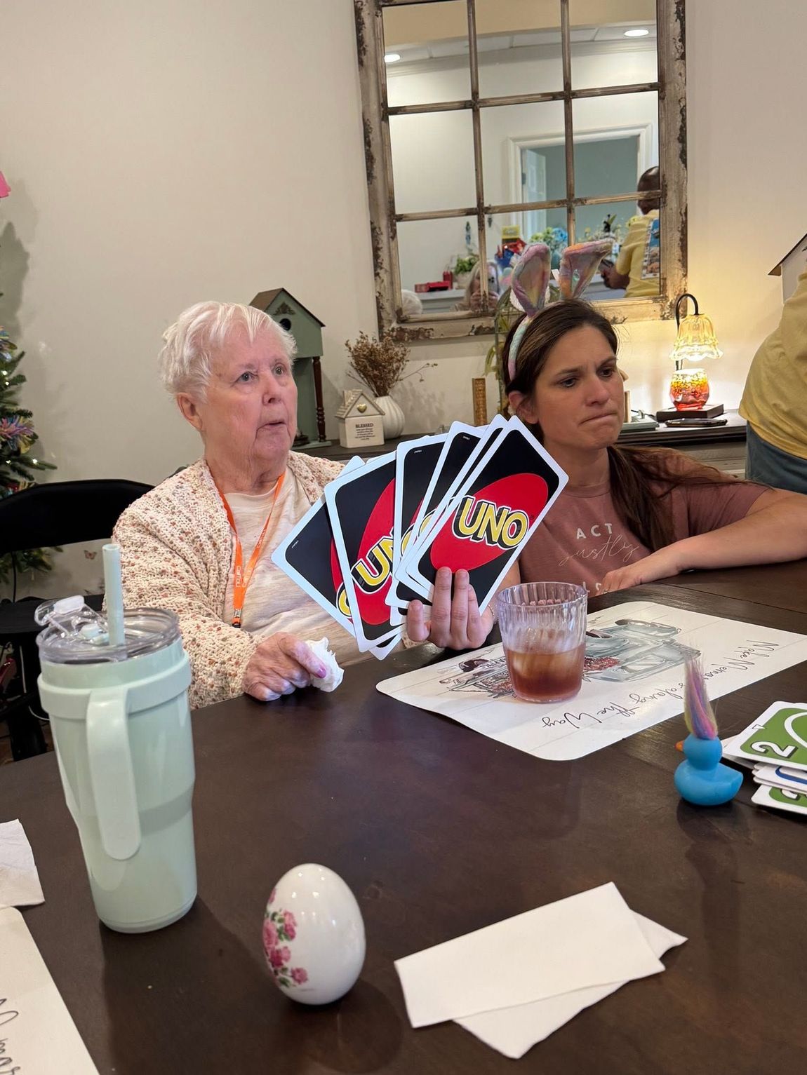 Two people at a table holding fan-shaped playing cards, with drinks and papers in a cozy dining room.