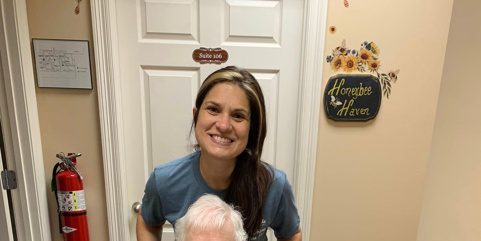 A person with long hair smiles behind an elderly individual; a fire extinguisher is on the wall near a door.