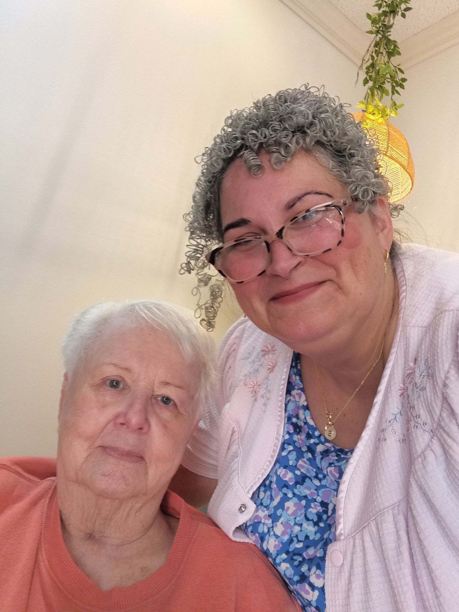 A selfie of two people smiling together indoors; one wears an orange sweater, the other wears a blue floral top and cardigan.