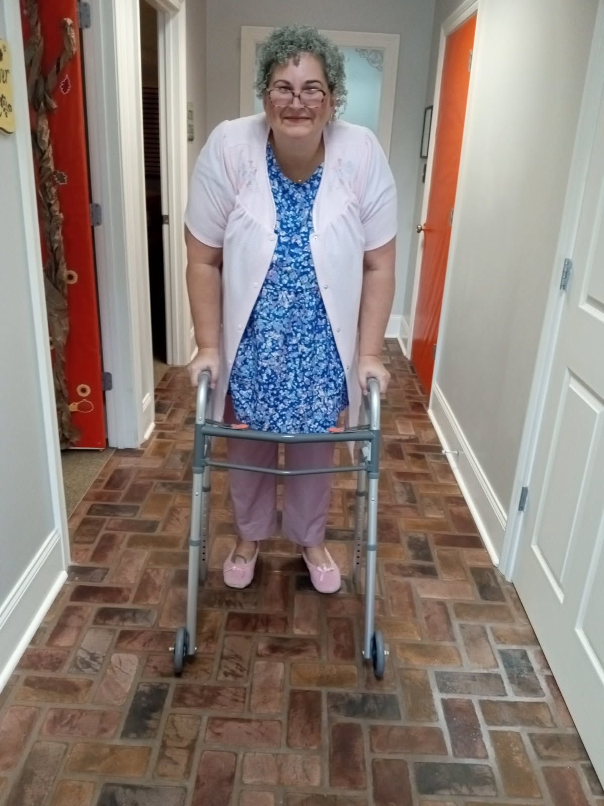 A person in a blue patterned top and pink cardigan stands in a brick-floored hallway, using a metal walker for support.