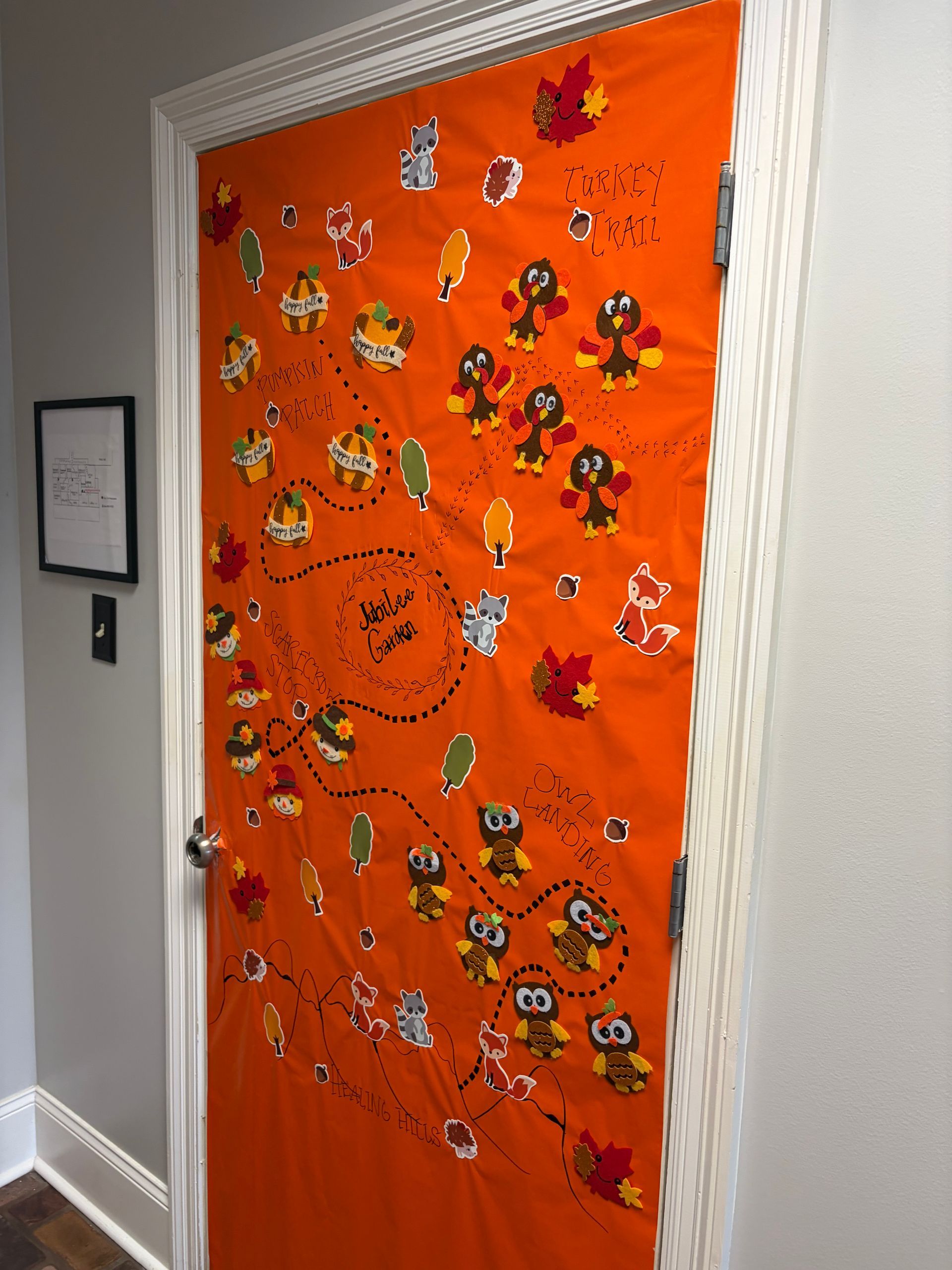An orange door decorated with fall-themed paper cutouts of turkeys, owls, and leaves arranged along a dotted trail path.