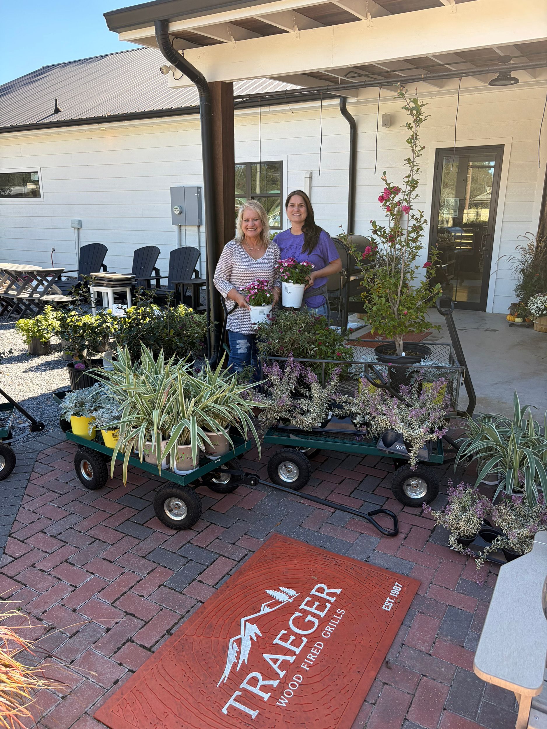 Two people standing with two wagons full of potted plants on a brick patio in front of a building with a Traeger mat.
