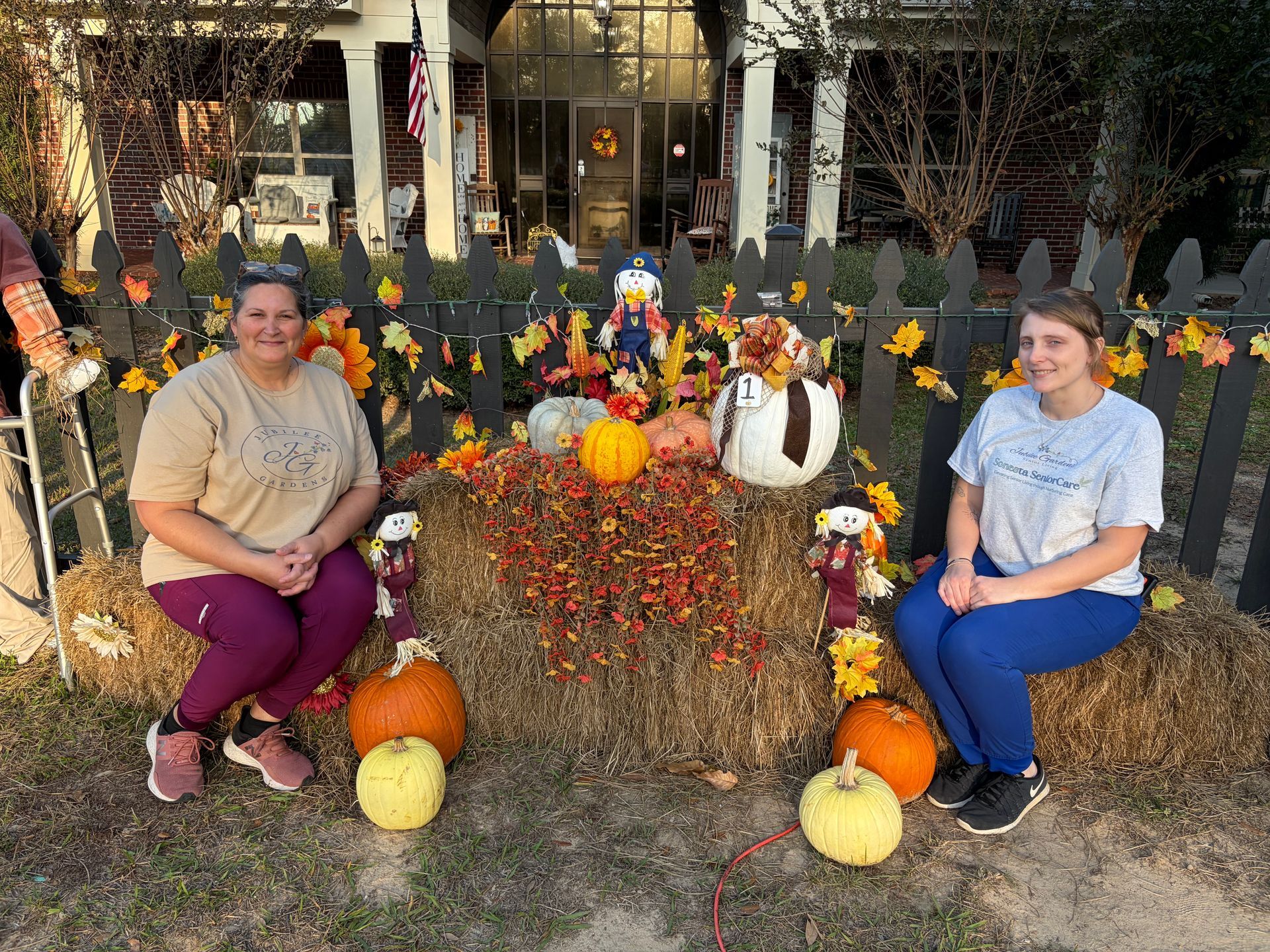 Two people sit on hay bales decorated with pumpkins and autumn leaves in front of a house with a picket fence.