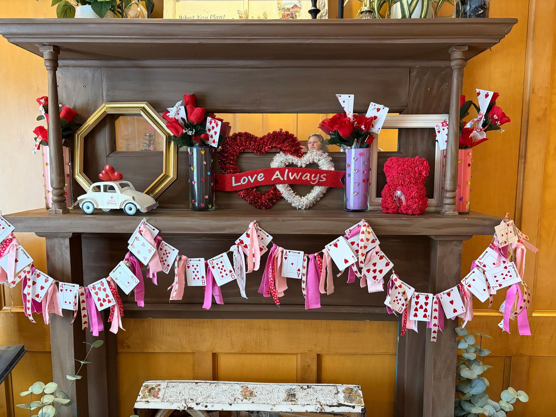 A mantel decorated for Valentine's Day with heart-themed decor, playing cards, flowers, and a banner of cards and ribbons.