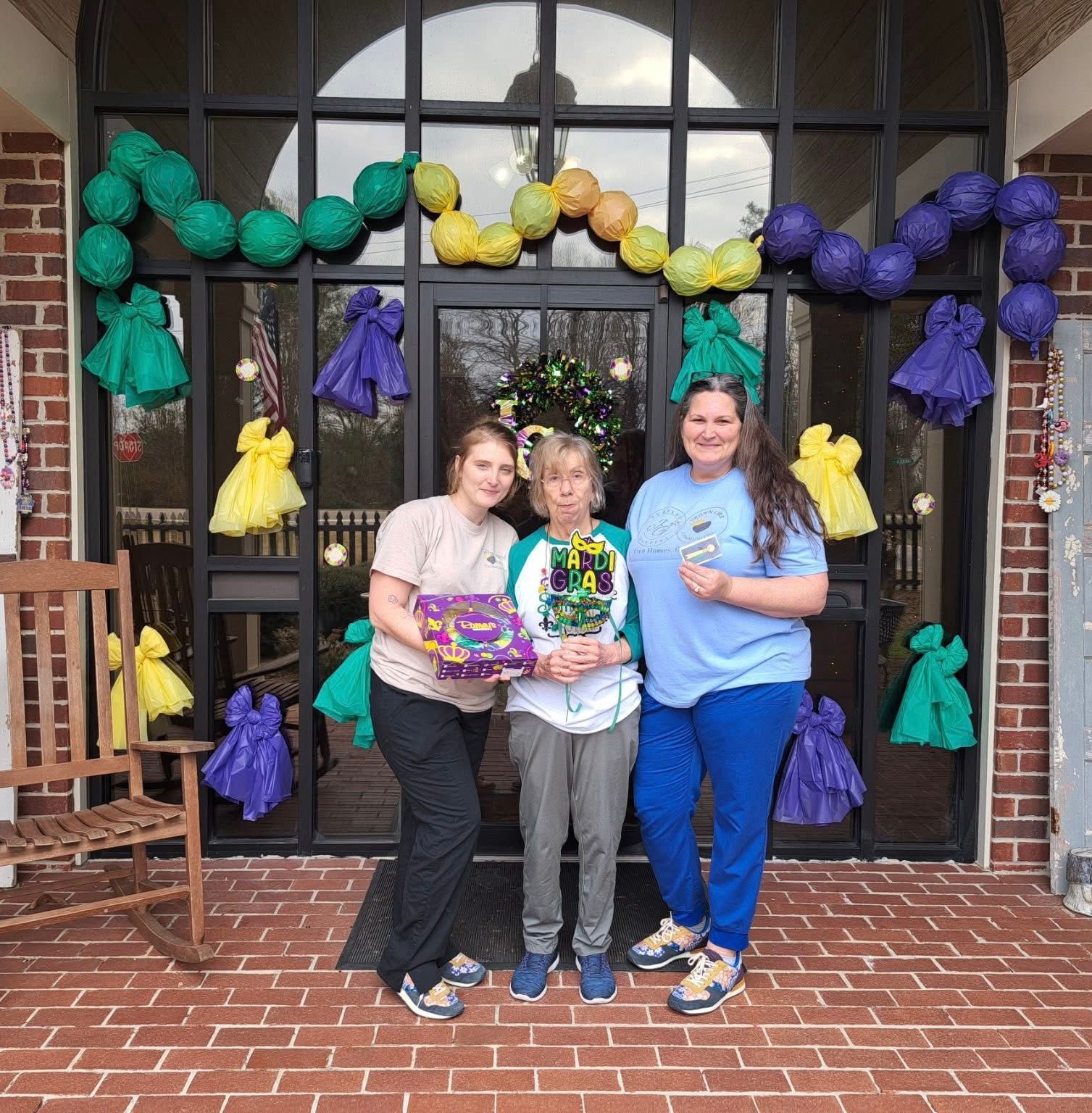 Three people stand in front of a glass door decorated with green, yellow, and purple garland for Mardi Gras.