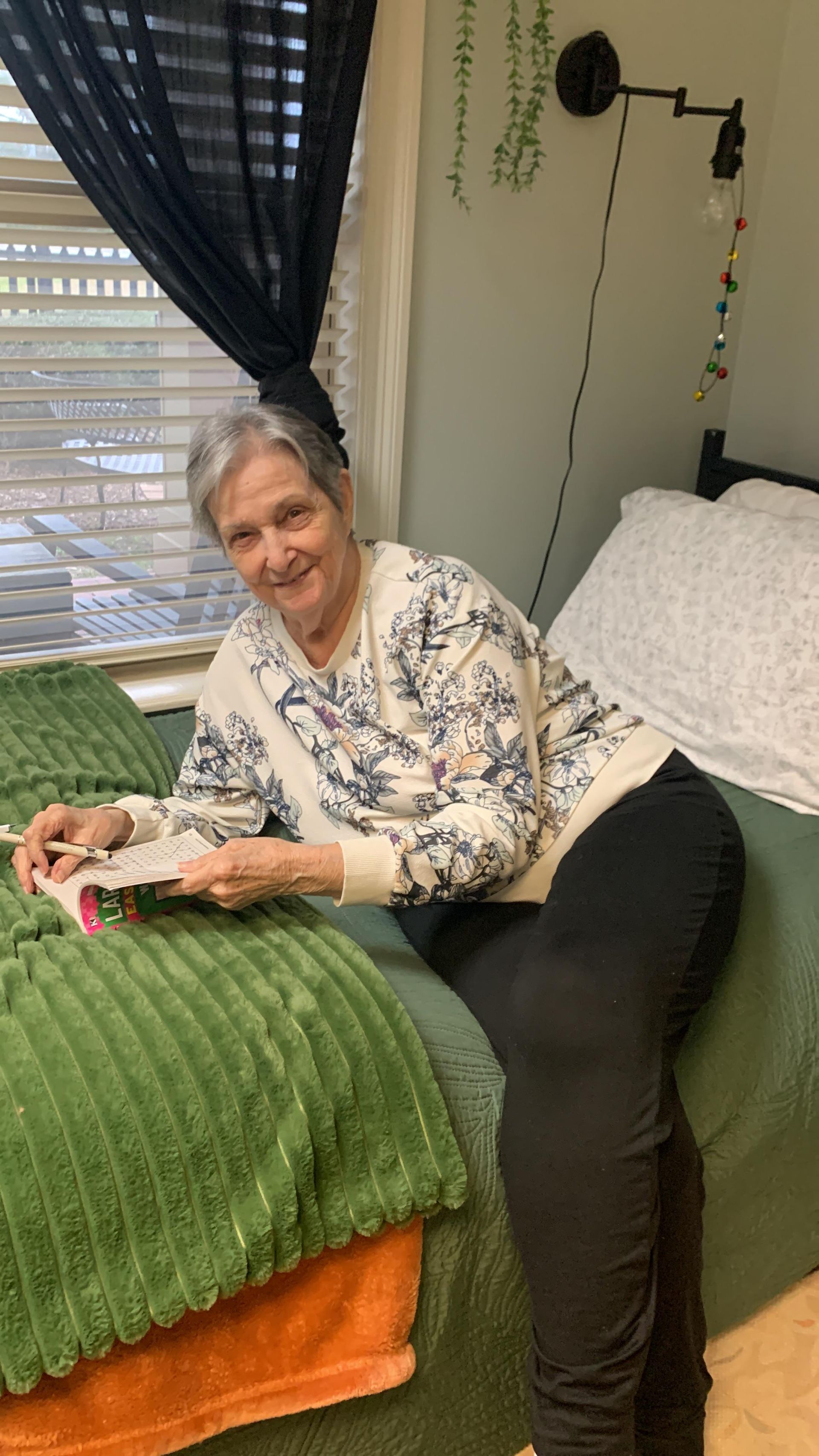 A person sits on a bed near a window, smiling while working on a needlework project.