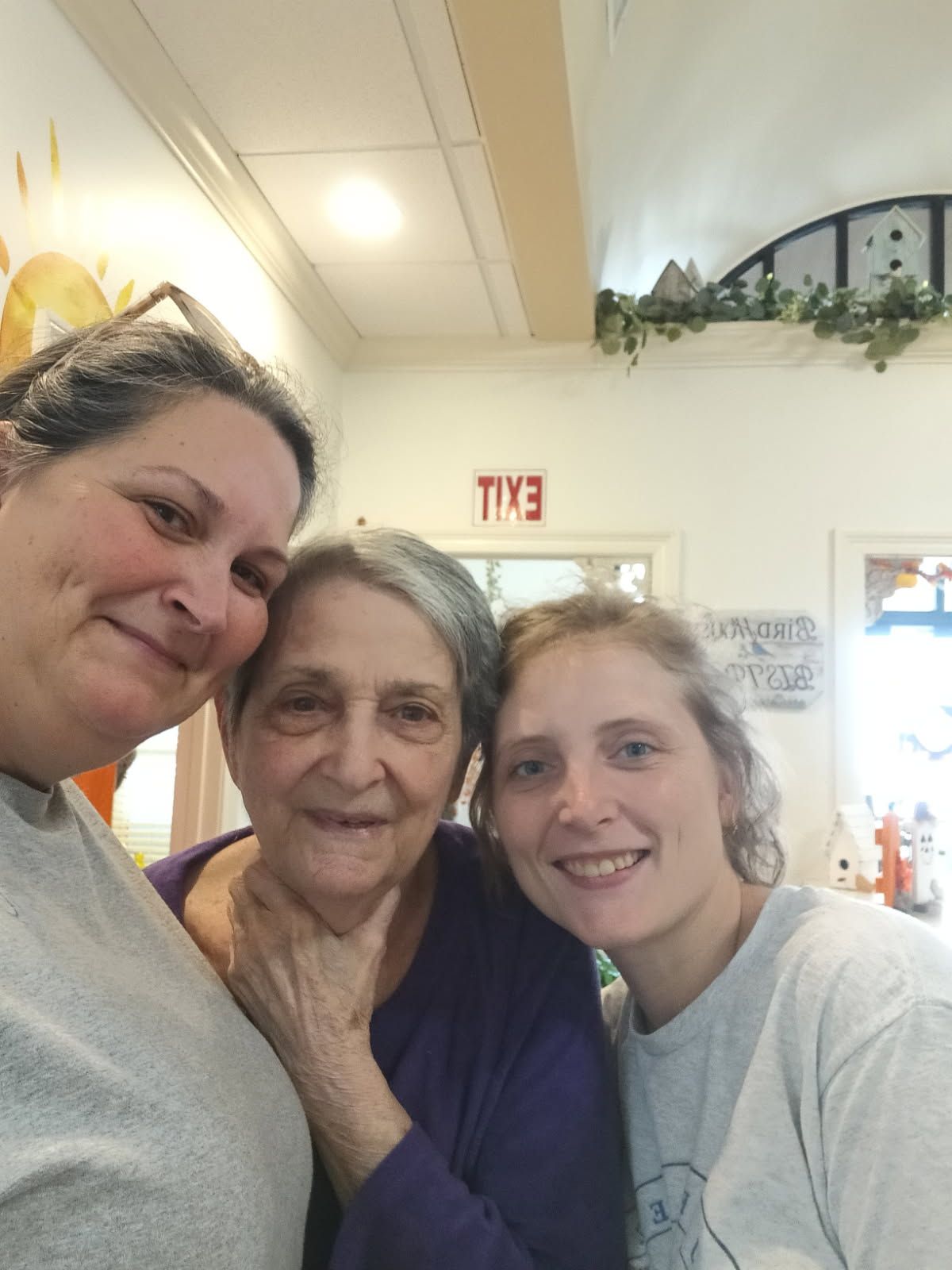 Three people smiling together in a brightly lit indoor space with an exit sign on the wall behind them.