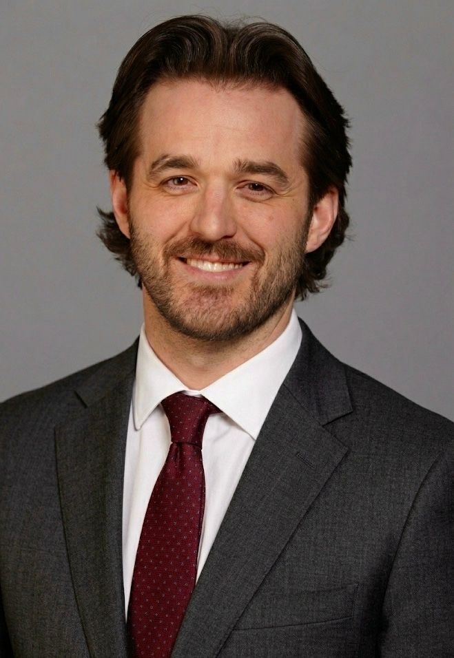 Man in black shirt, looking at the camera, against a grey backdrop.