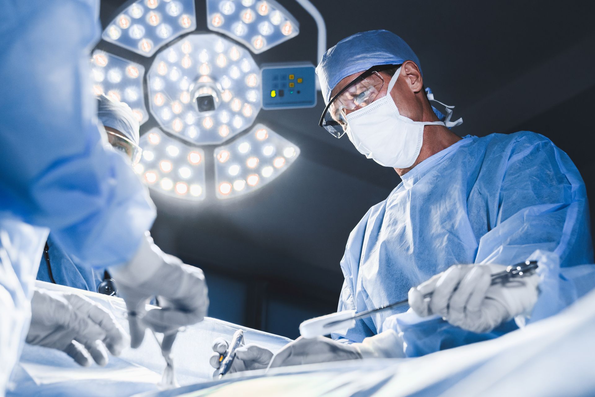 Surgeon in scrubs, mask, and glasses operating under bright surgical lights.