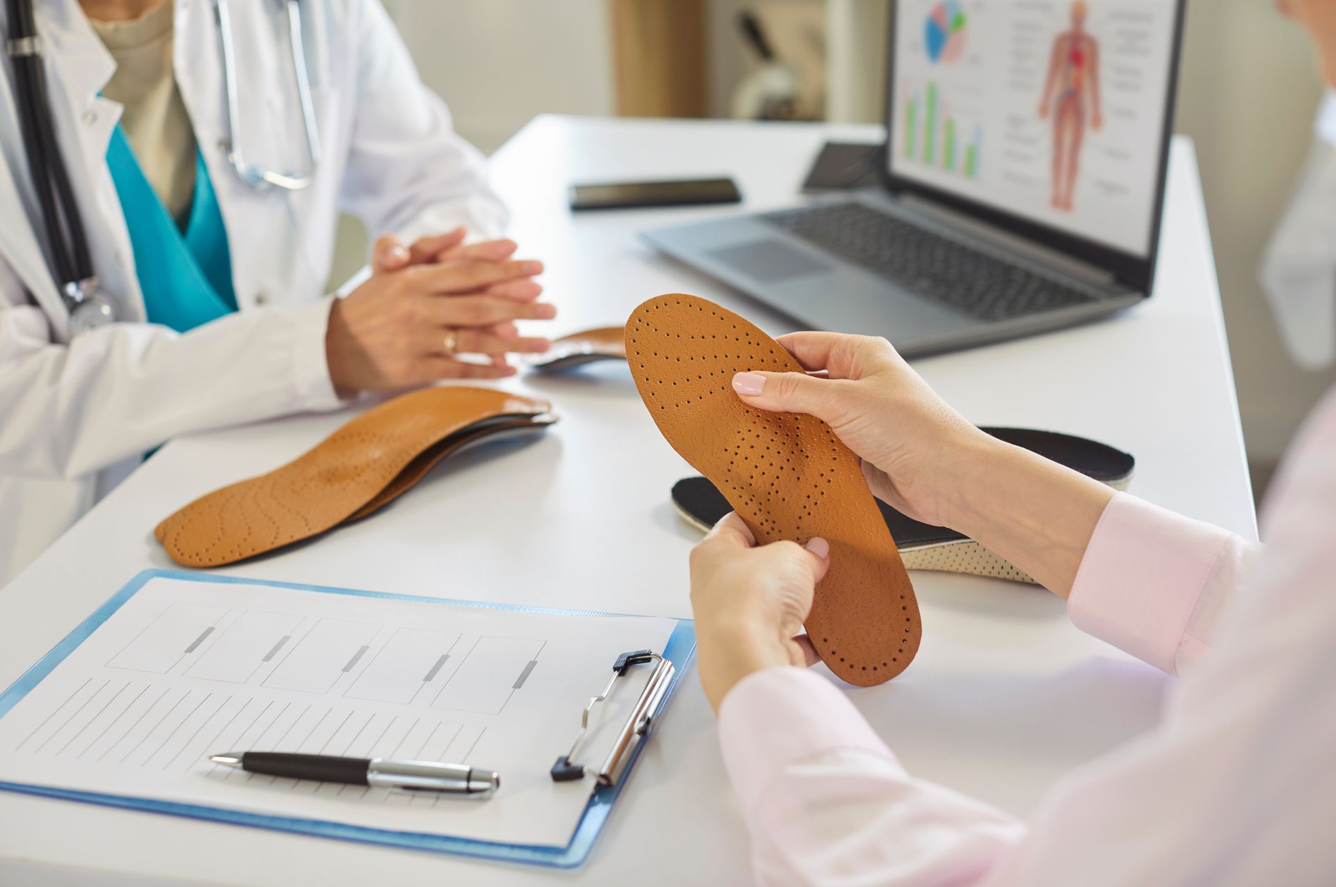 Doctor showing insoles to a patient at a desk with a laptop and clipboard.