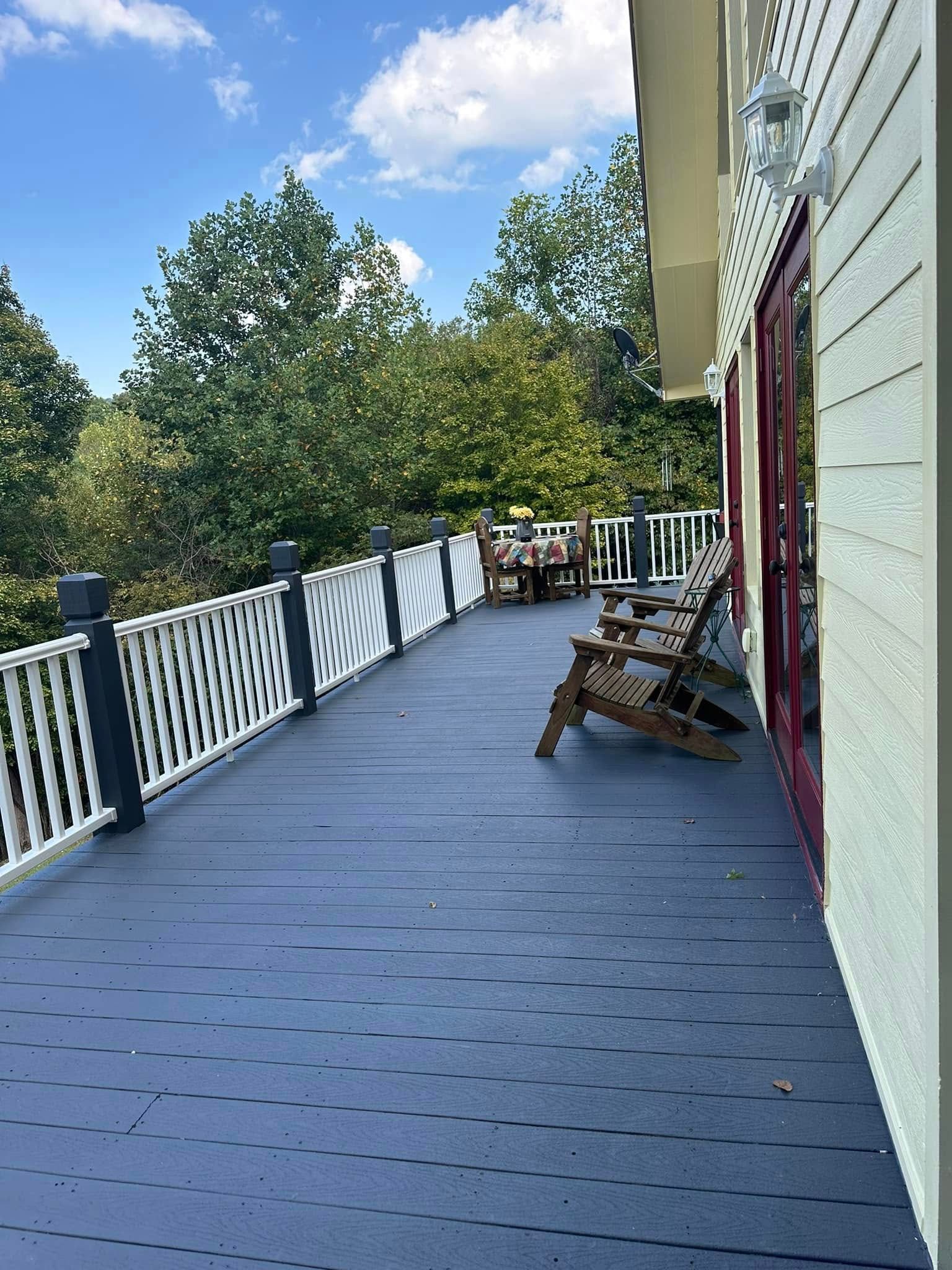 Deck with blue floor and white railing, two wooden chairs, and table, overlooking trees.
