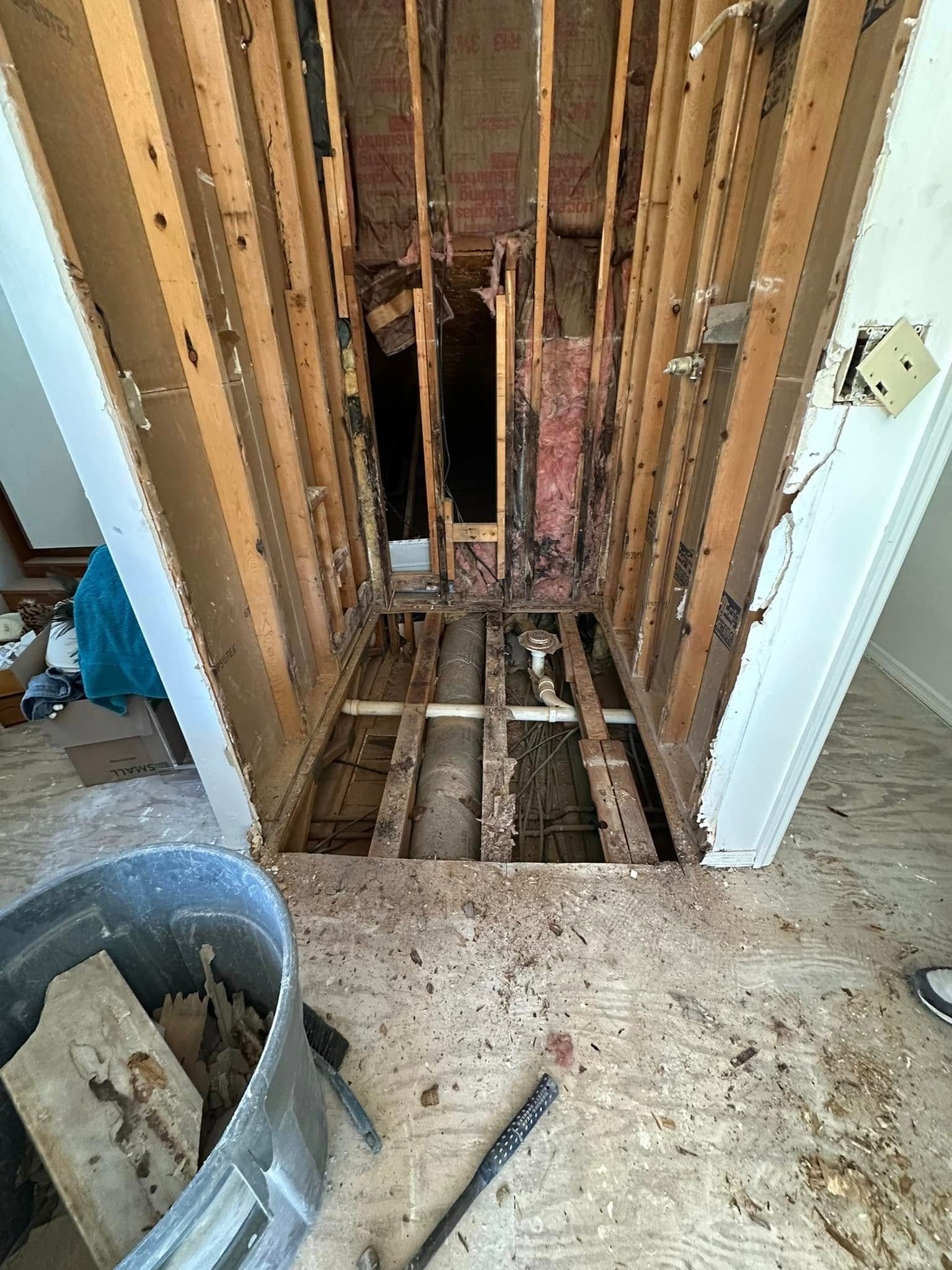 Interior view of a damaged hallway with exposed framing and missing flooring. Debris and a trash can are visible.