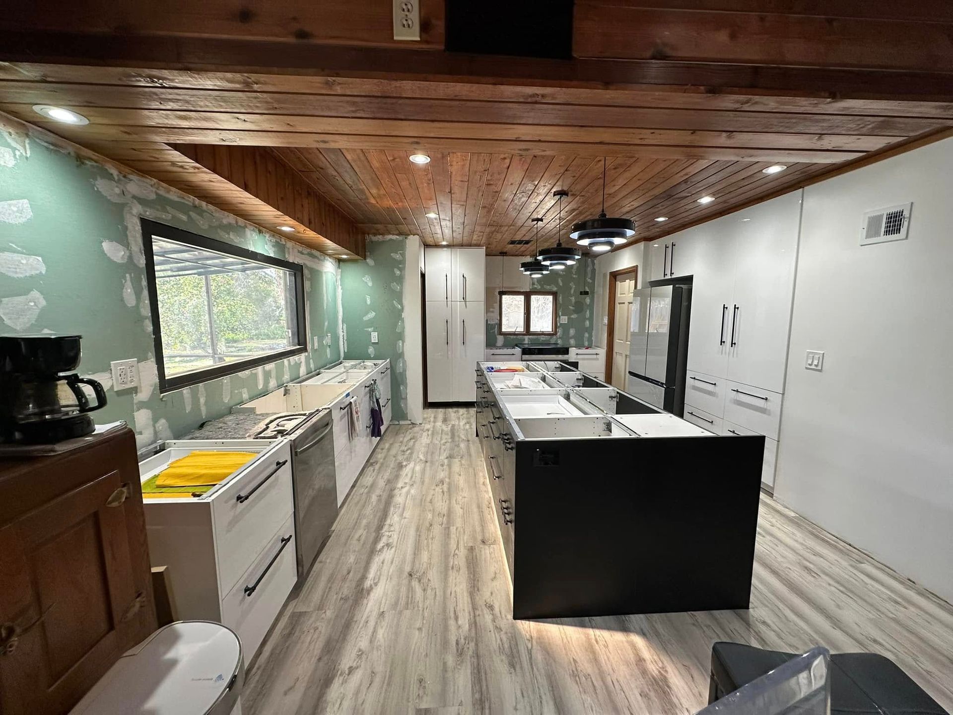Kitchen under renovation with light wood ceiling, white cabinets, black island, and light gray floor.