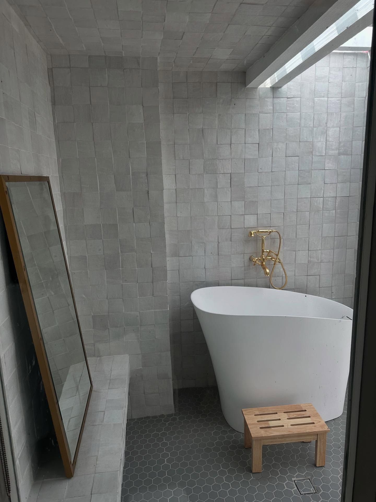 A modern bathroom featuring a white tub, gold fixtures, textured gray tile walls, and a wooden stool.