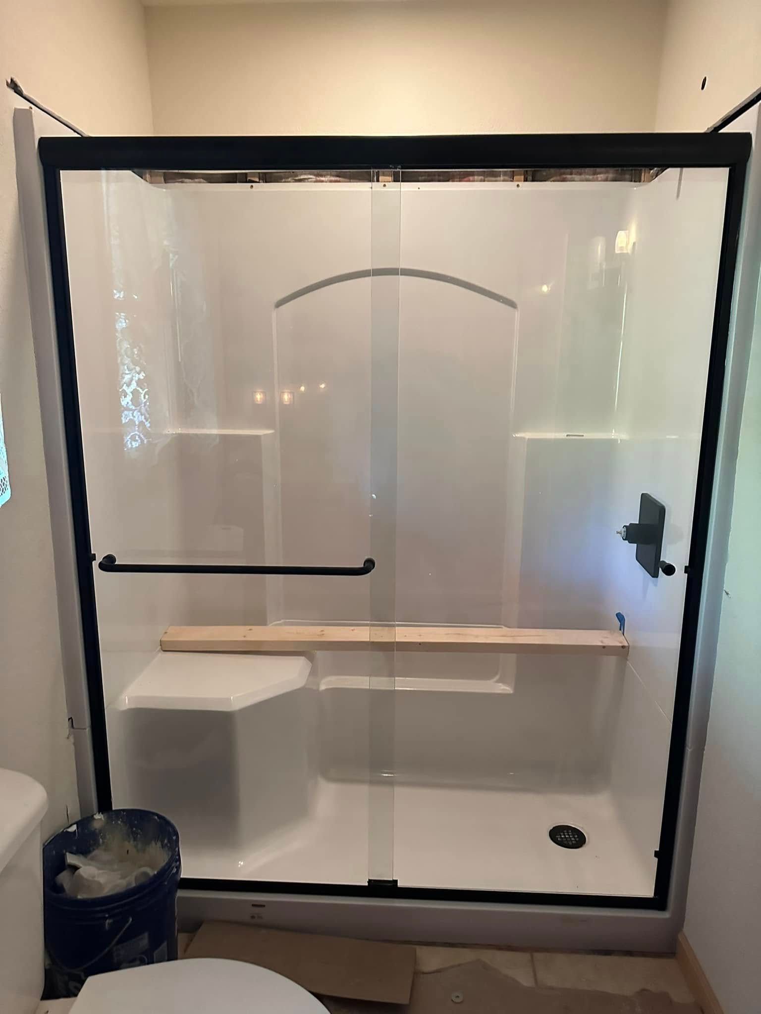 Black-framed shower stall with white walls, built-in seat, and shelf, next to a toilet; construction supplies visible.