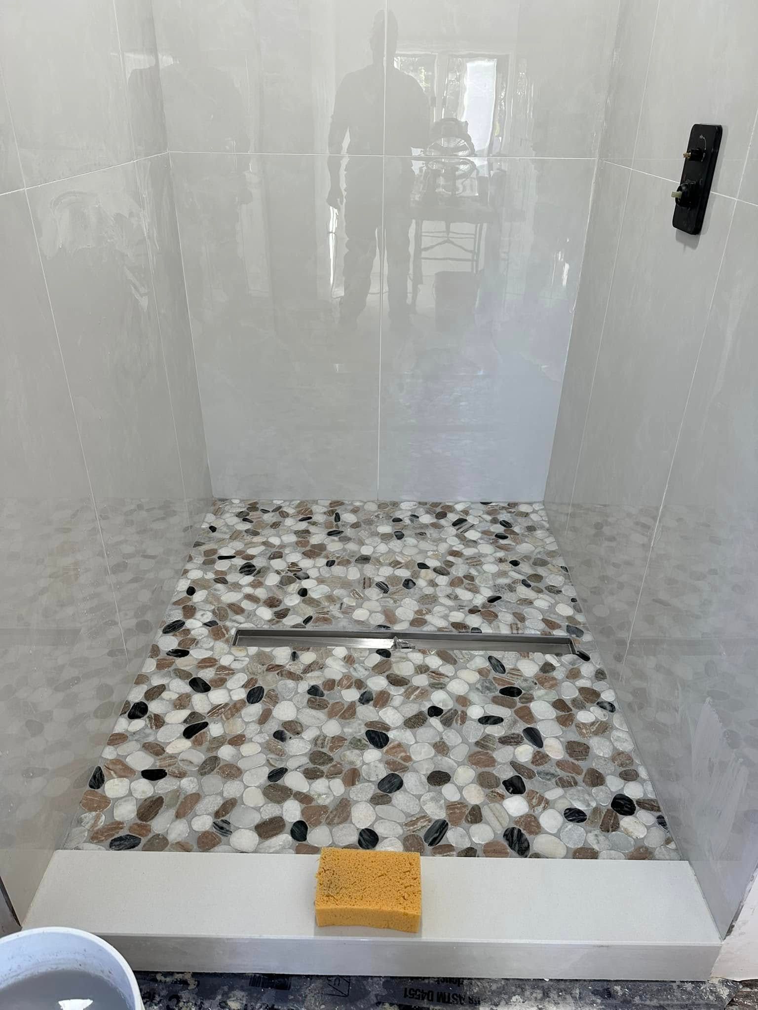 Shower with pebble tile floor and linear drain, white tiled walls. A sponge sits on the curb.