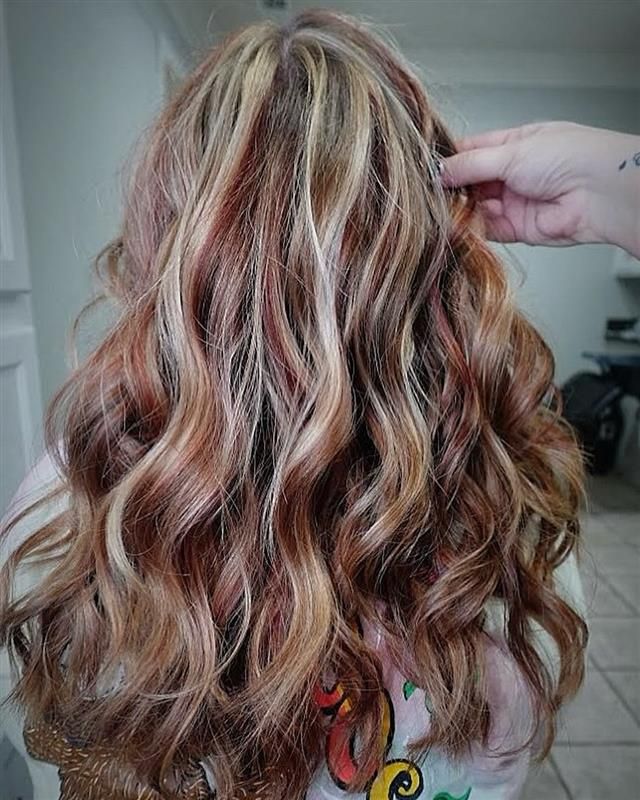 Smiling person with wavy blonde hair, looking down with a hand on the chest, against a neutral background.