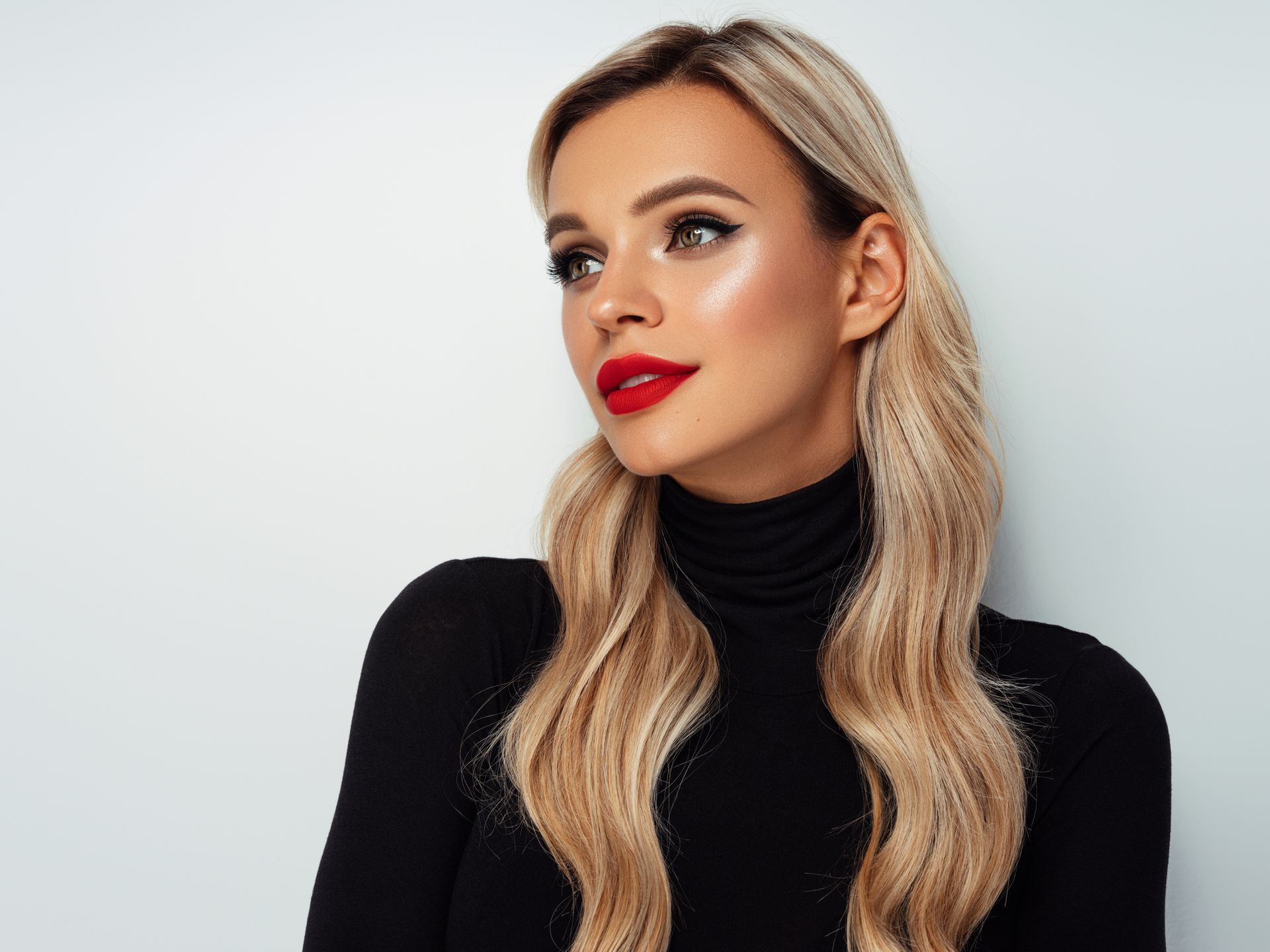Woman with blonde wavy hair, red lipstick, wearing black turtleneck, looking to the side.