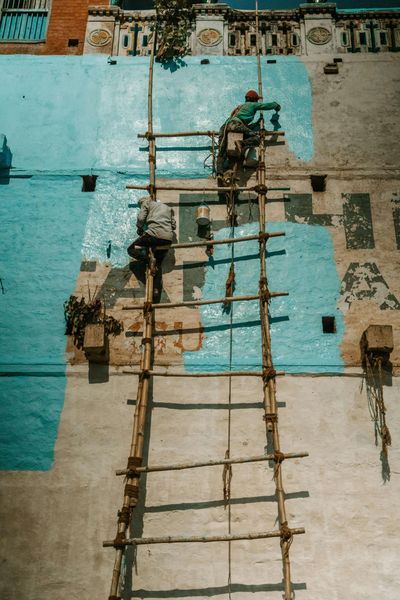 Two people painting a blue wall, using a bamboo ladder.