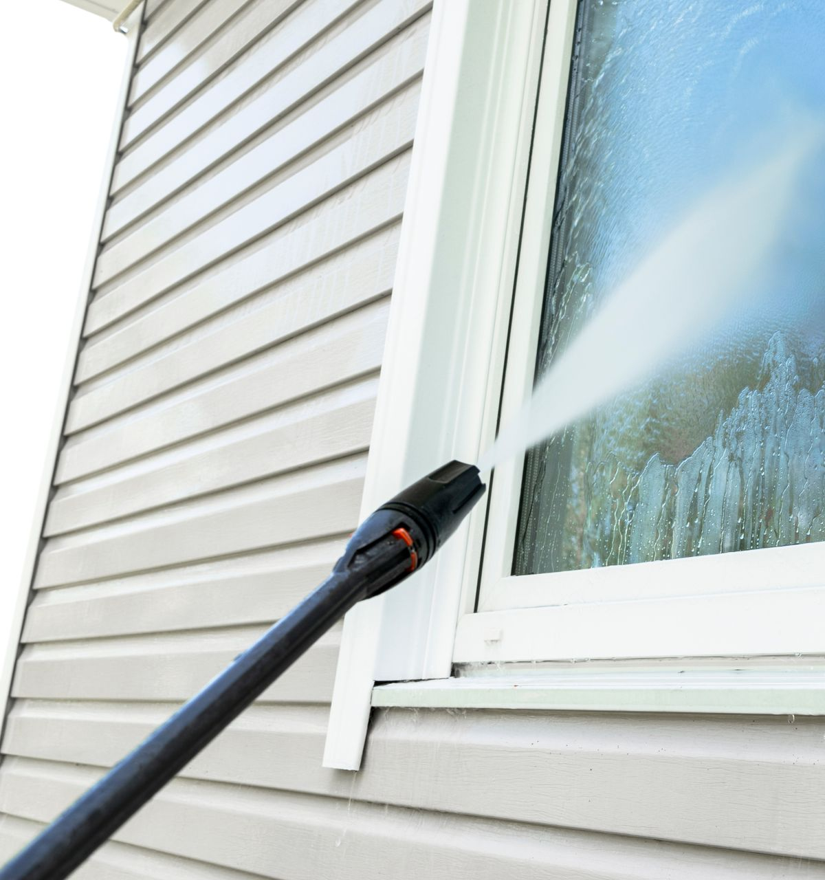 Pressure washer cleaning a window on a house with gray siding.
