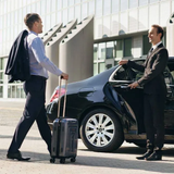 Man exiting black car with luggage; driver holds open the door outside modern building.