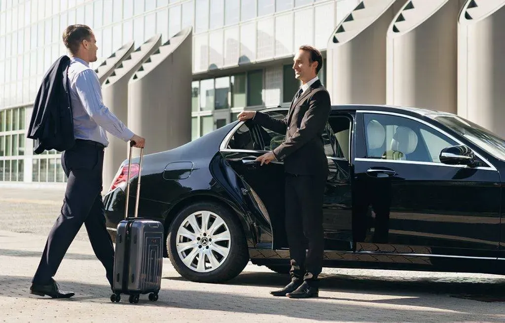 Man with suitcase approaches black car as attendant opens door. Building in background.