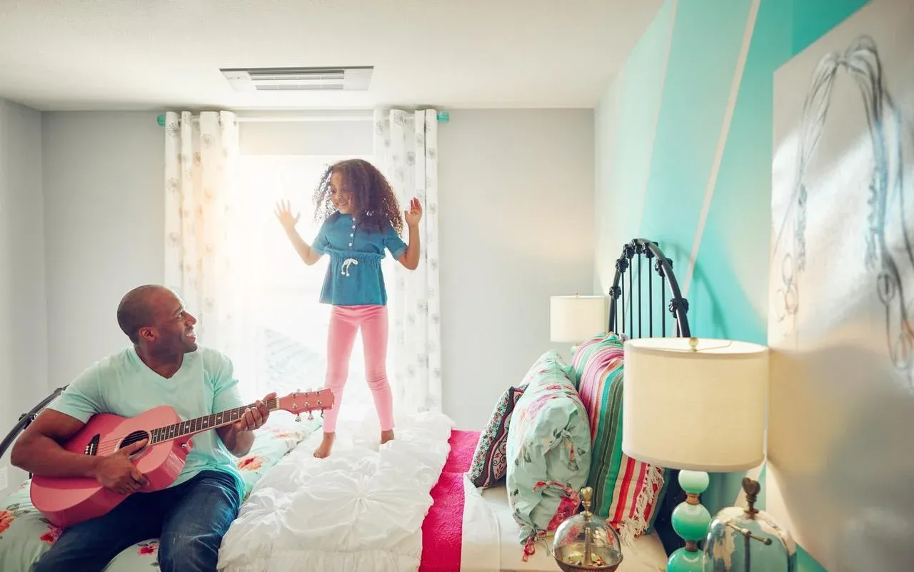 A man is playing a guitar while a little girl is jumping on a bed.