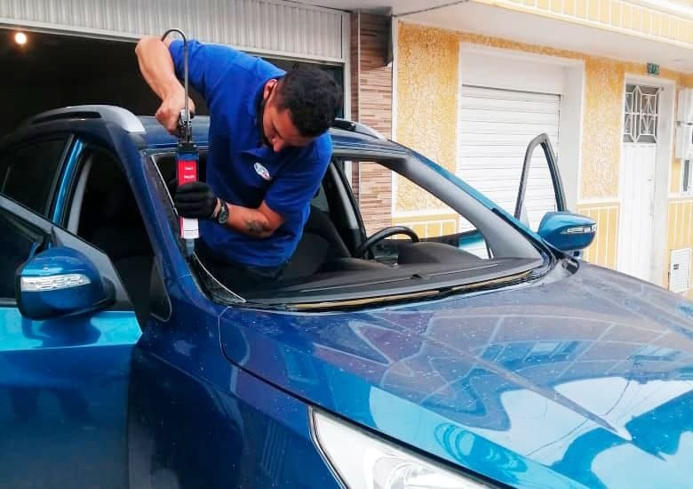 Hombre instalando un parabrisas en un coche azul, trabajando en un taller mecánico.
