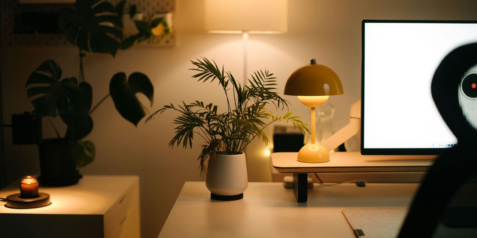 A warmly lit workspace with plants, a desk lamp, and a shadowy figure with glowing eyes.