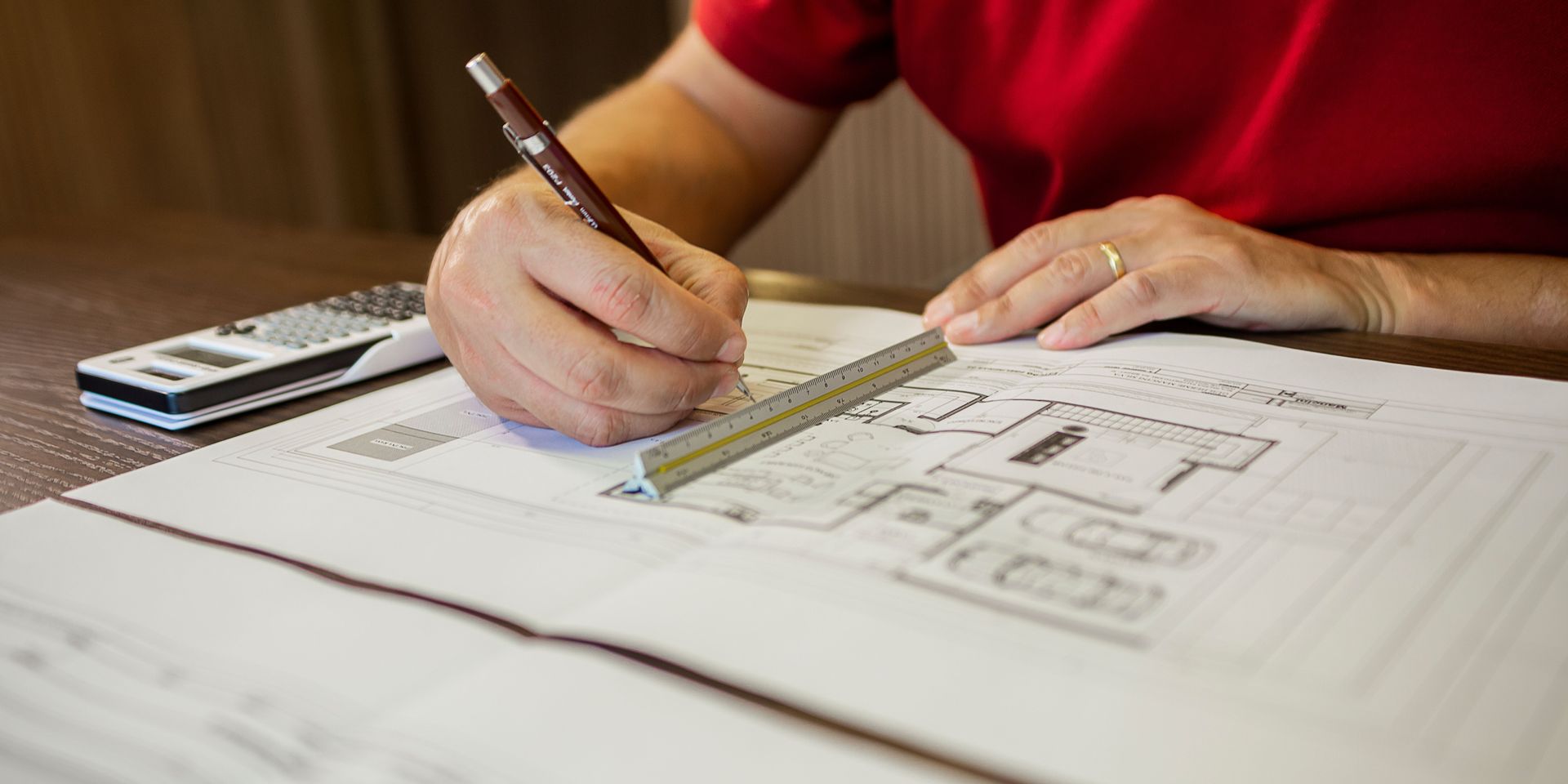 Person in red shirt drawing with pencil on blueprints.