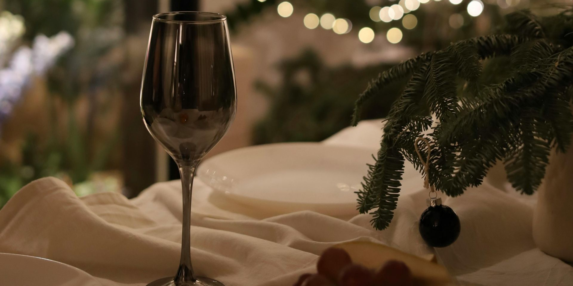 A champagne glass sits on a white tablecloth, decorated with fir branches and lights.