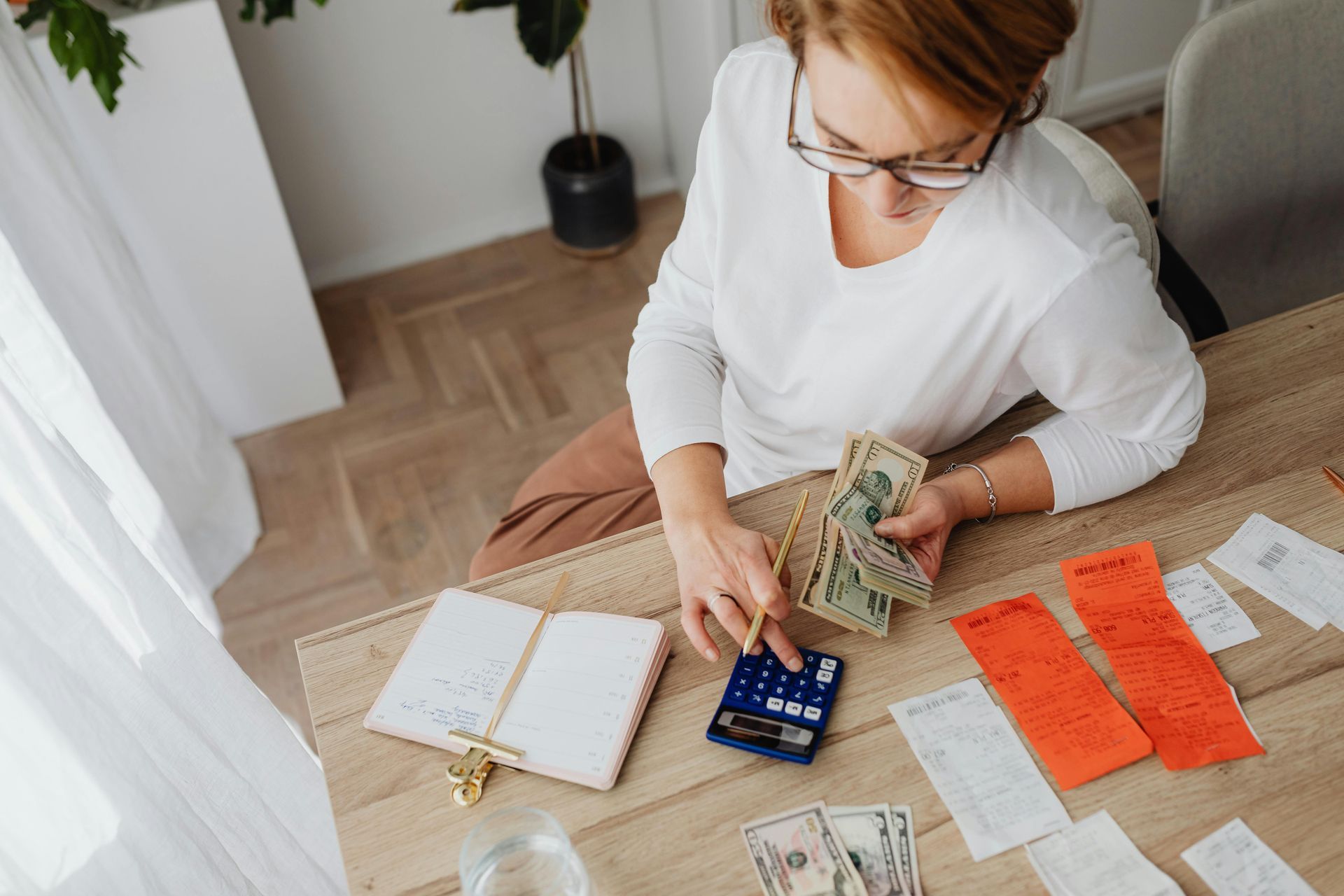 Woman counting money with a calculator, receipts, and a notebook on a wooden table.