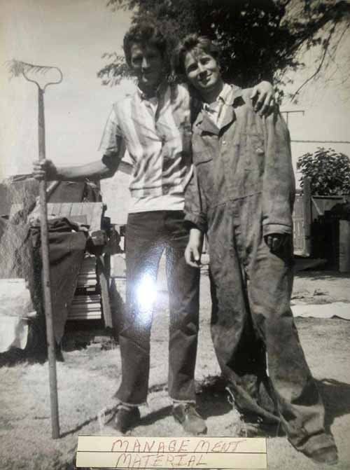 Two people posing. Man holds rake, woman in jumpsuit. Outdoors, text reads Two people posing. Man holds rake, woman in jumpsuit. Outdoors, text reads
