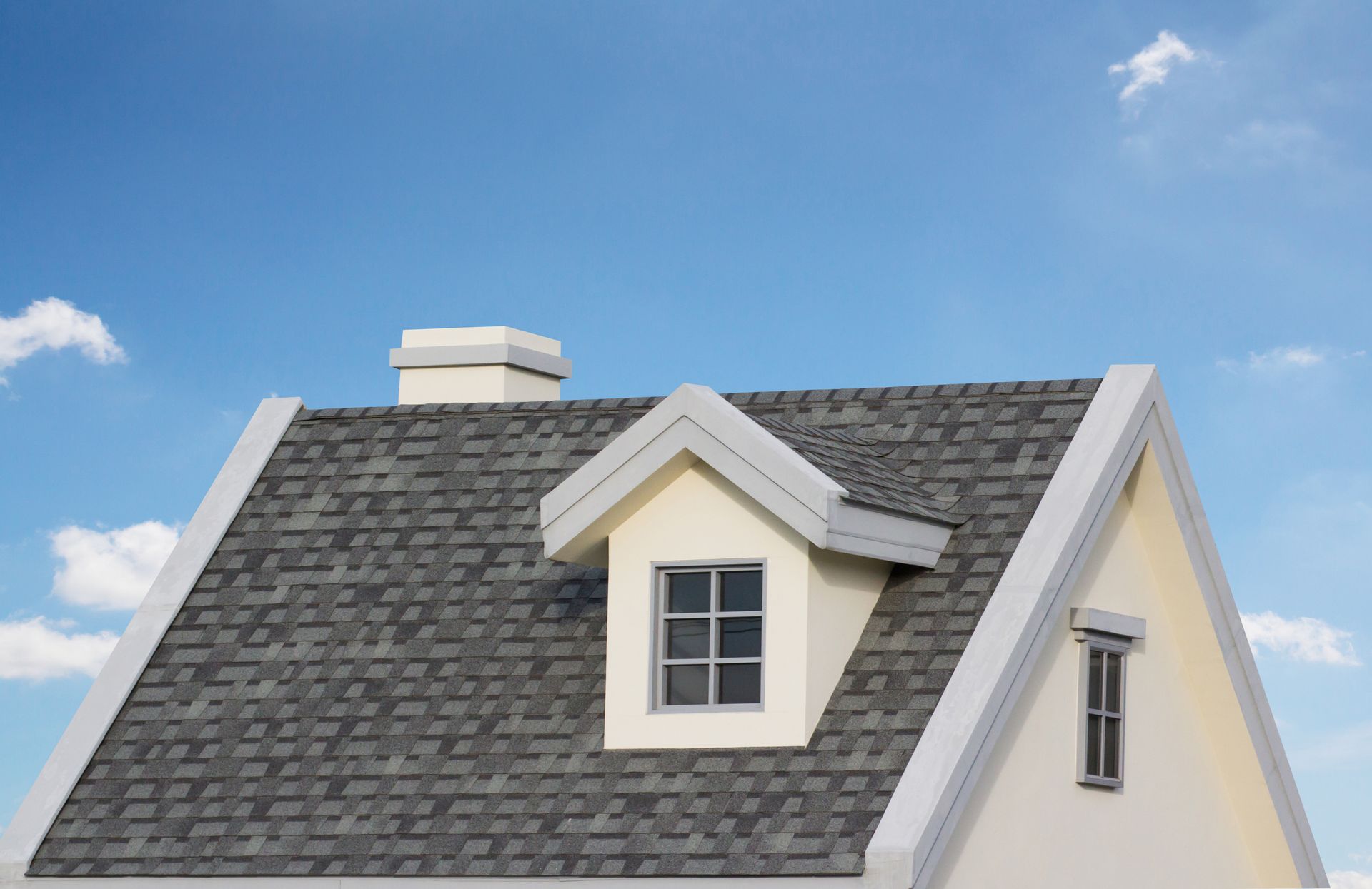 A house roof with gray shingles, a dormer window, and a chimney against a blue sky.
