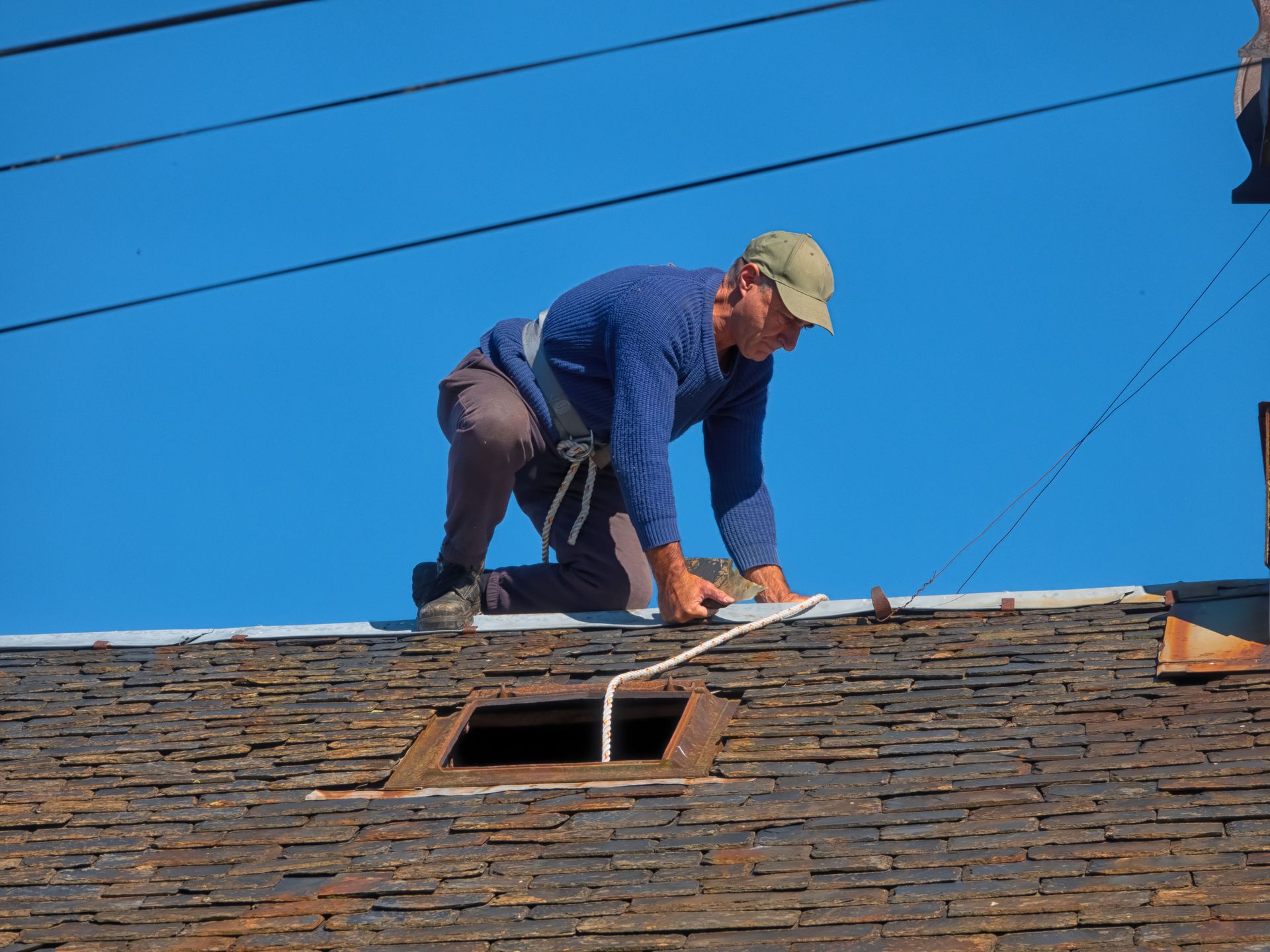 Man kneeling on a dark shingle roof, working; blue sky background.