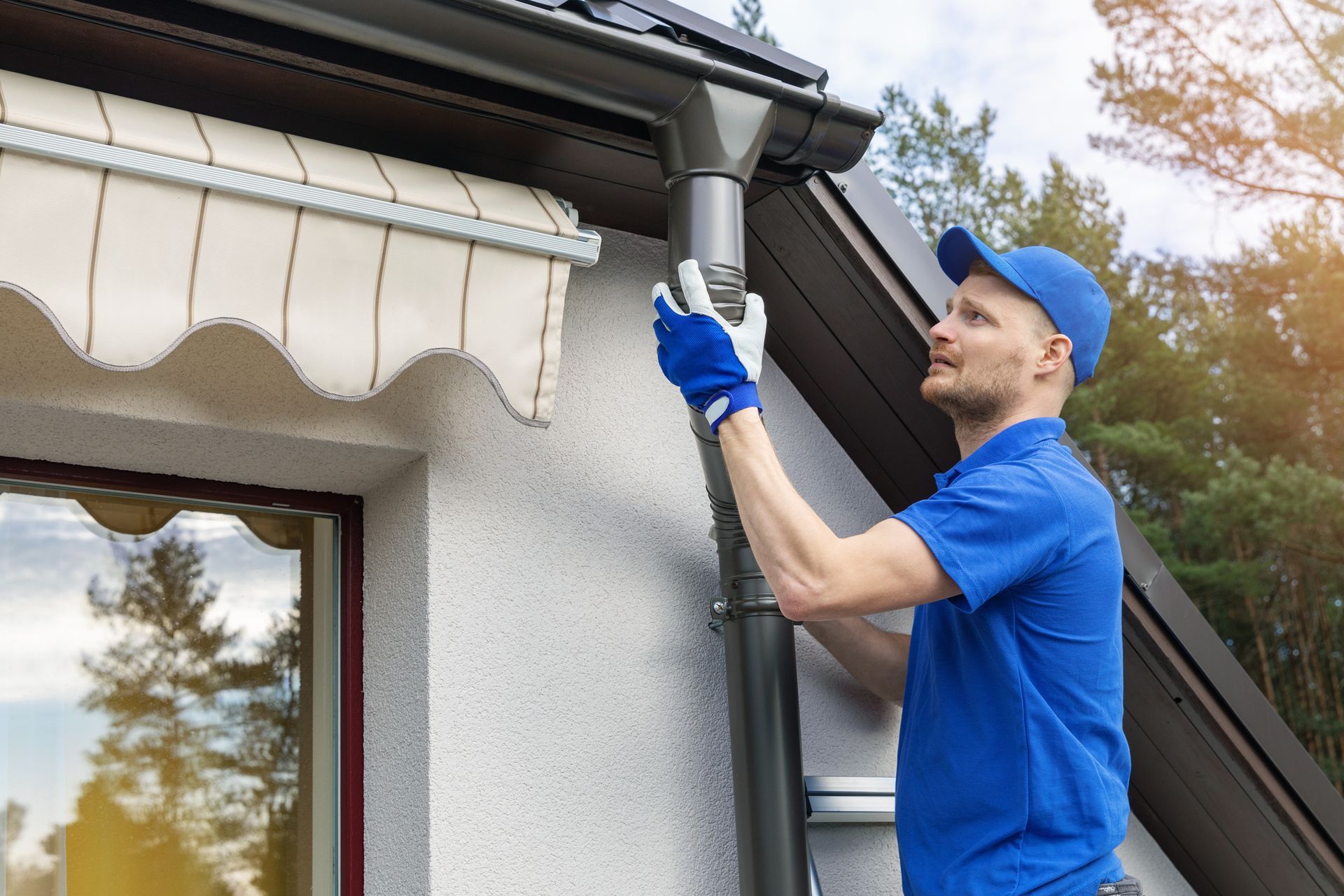 Man in blue installing a gutter downspout. White house, awning, and trees in the background.