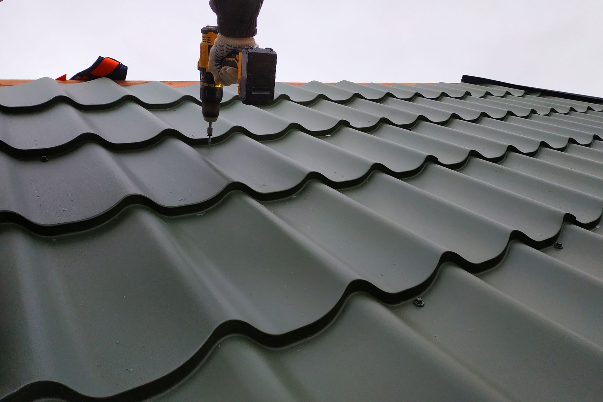 A person using a drill to attach green metal roofing panels on a rooftop.
