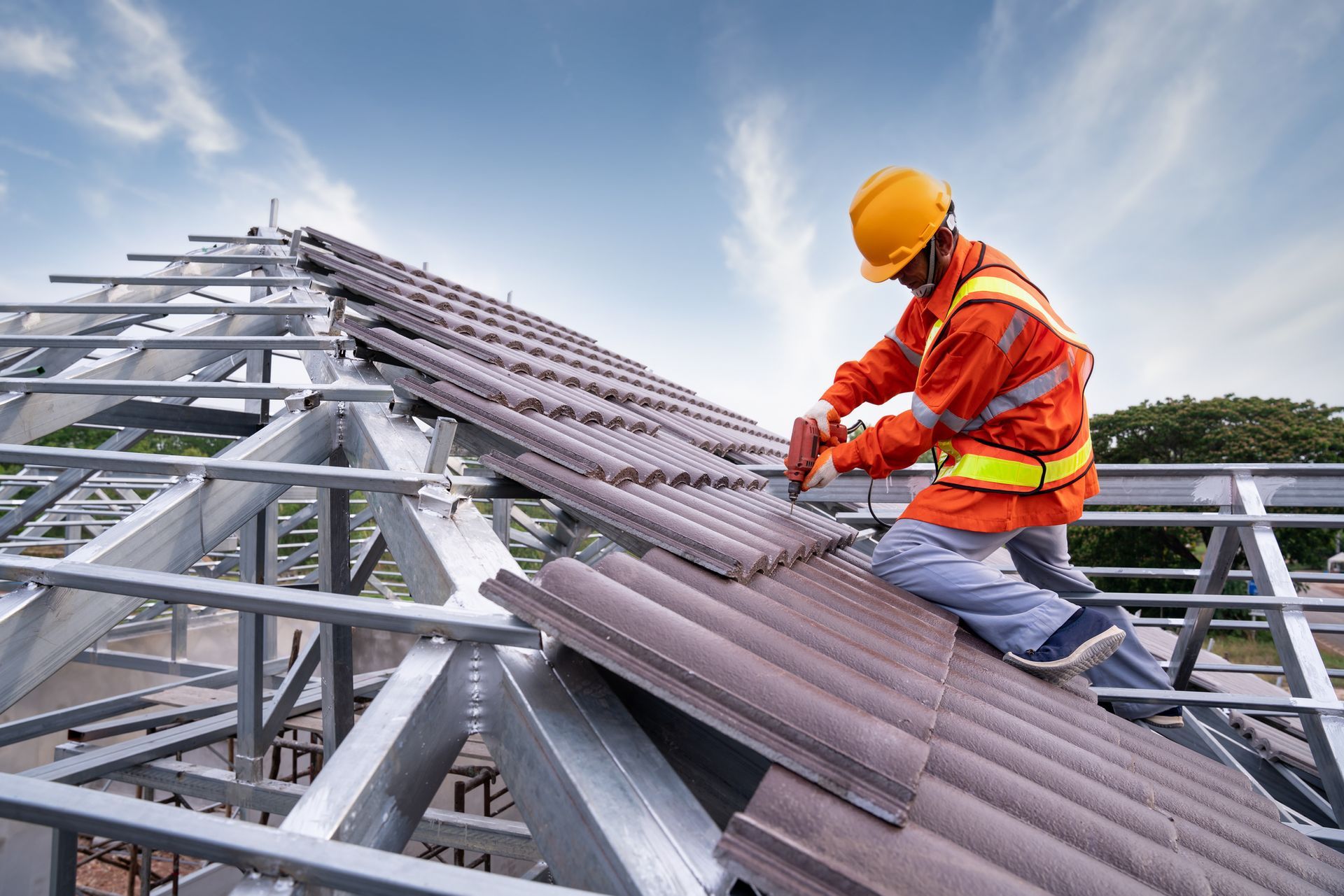 Roofer in orange safety gear installing tiles on a metal roof frame under a cloudy sky.
