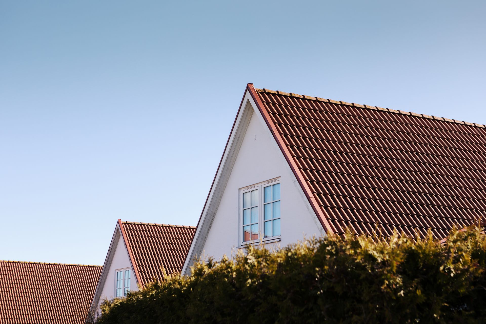 White houses with brown tile roofs against a blue sky, partially obscured by green shrubs.