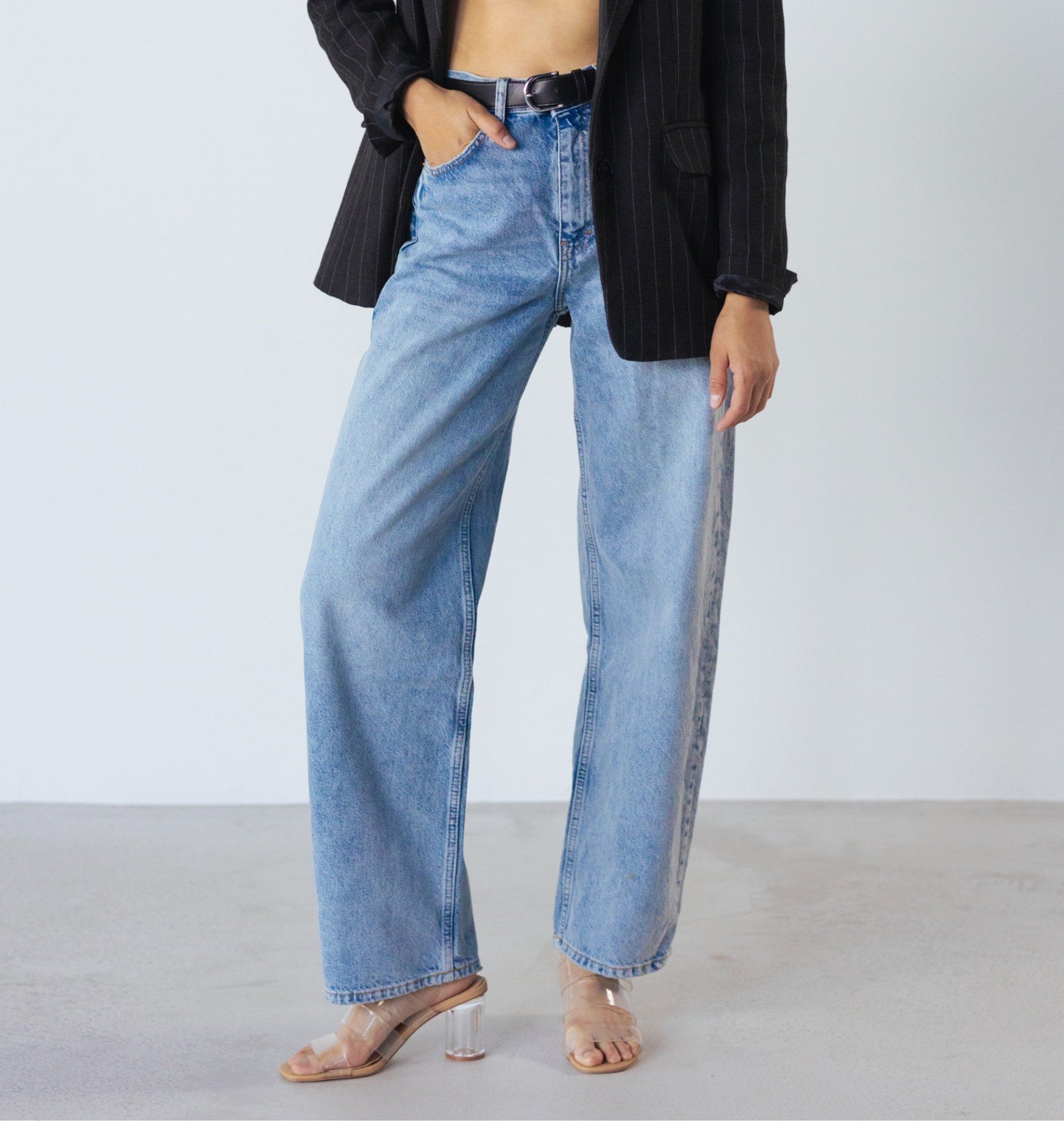 Person wearing light blue jeans and heels with a black blazer, standing against a plain wall.