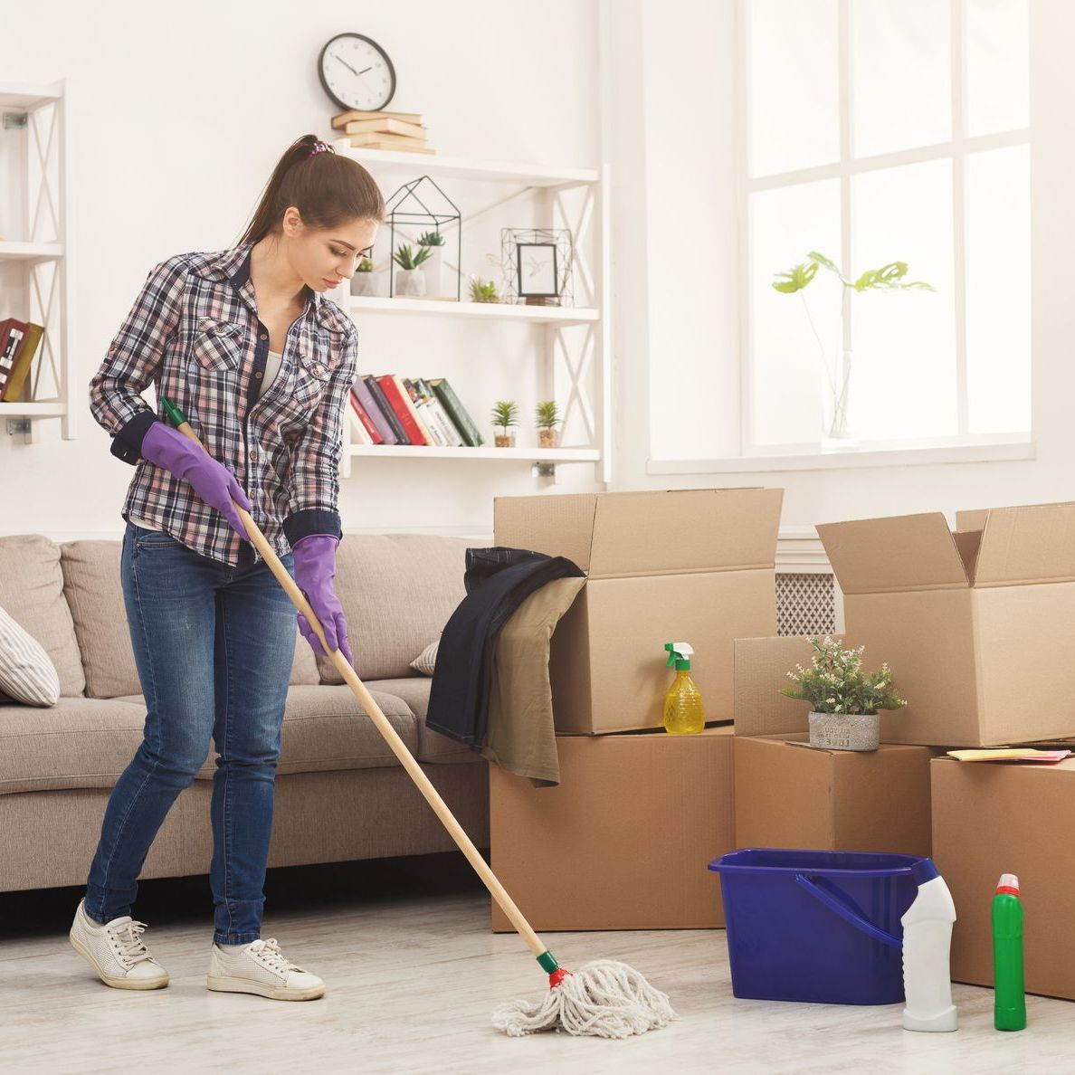 A woman is mopping the floor in a living room filled with boxes.