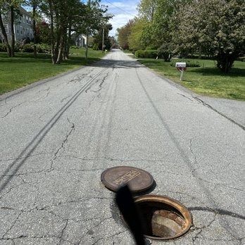 Open manhole in the cracked asphalt of a residential street with trees and houses in the background.